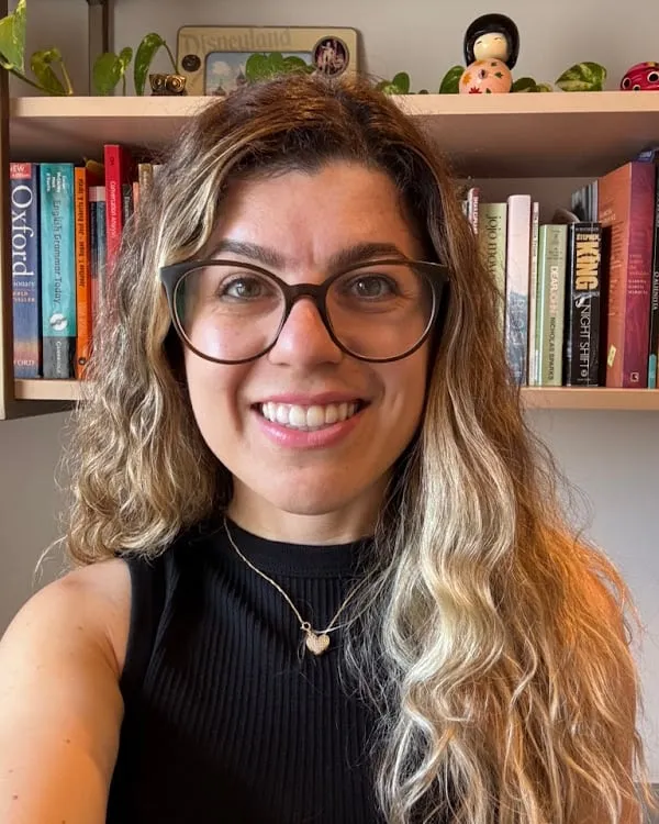 Camila Almeida sorrindo em frente à sua estante de livros de inglês