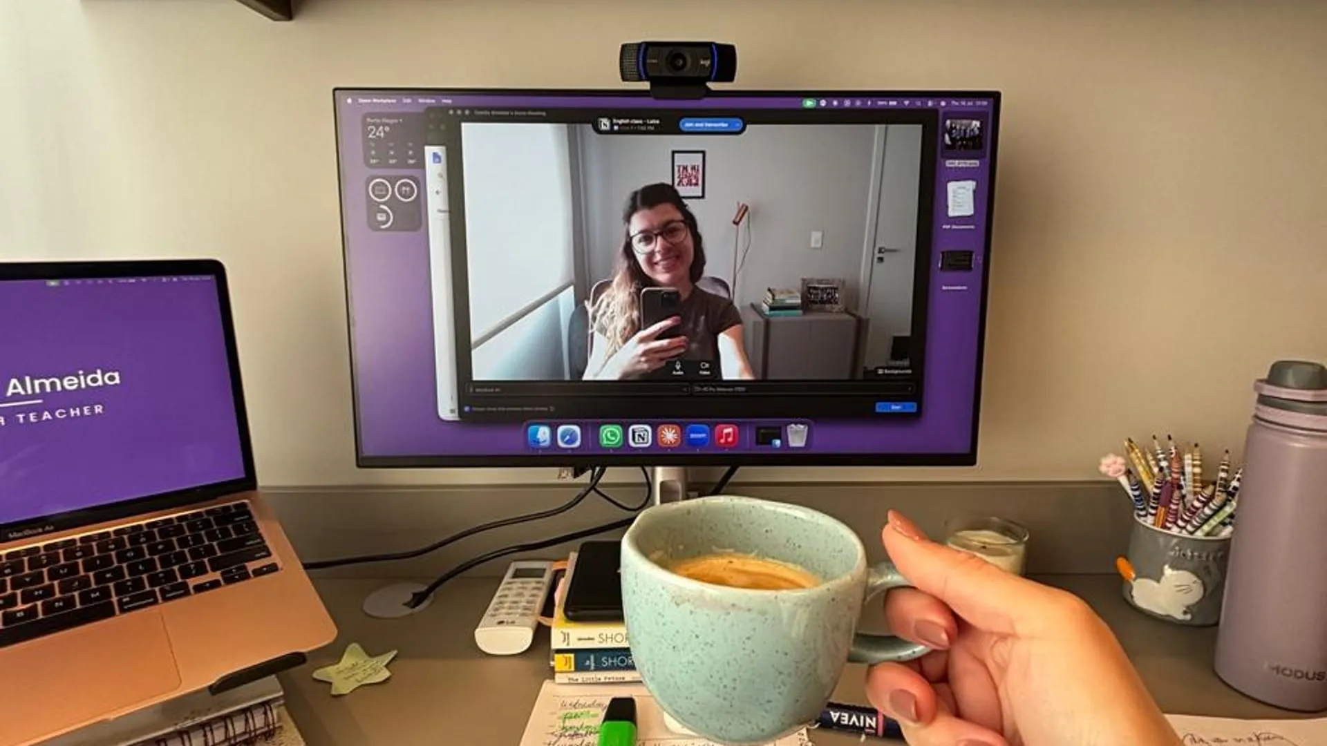 Mesa de trabalho da Camila: monitor com uma aula no Zoom, notebook e estante de livros ao fundo