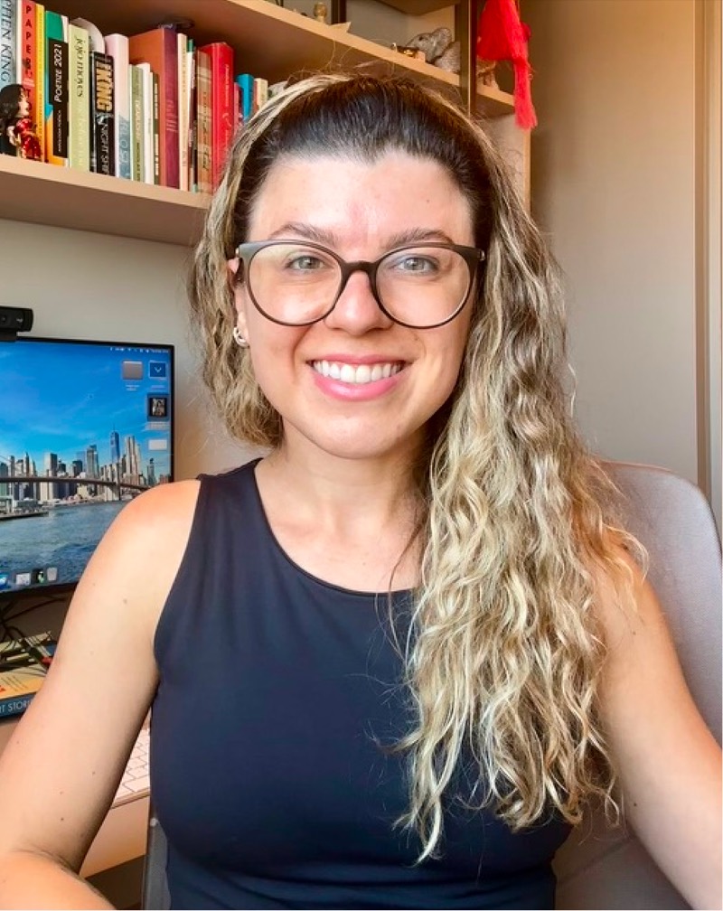 Camila Almeida sorrindo em frente à sua estante de livros de inglês